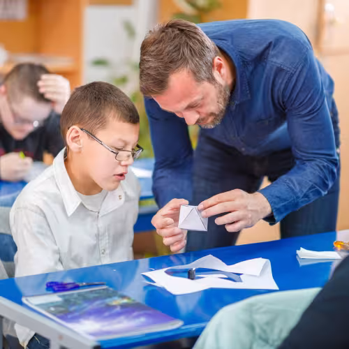 Ein Schulbegleiter hilft einem Schüler beim Falten einer geometrischen Form.